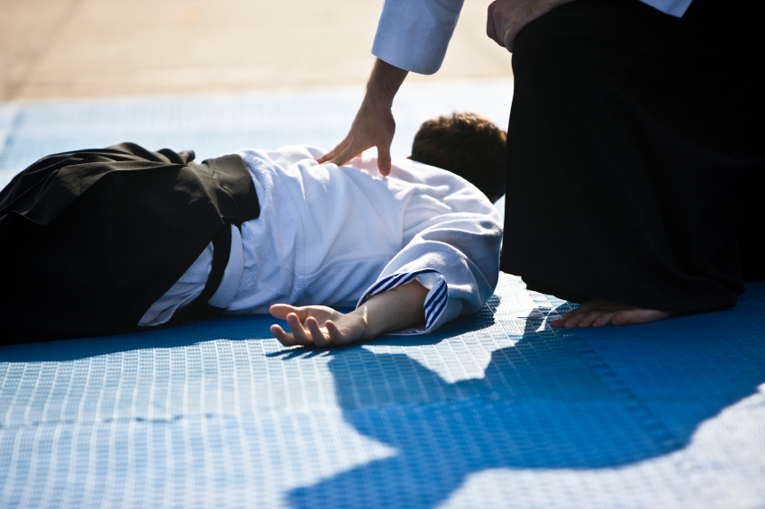 Eine Person in einem weißen Gi liegt auf einer Matte, während eine andere Person in einem schwarzen Hakama daneben kniet und eine Hand auf deren Rücken legt.