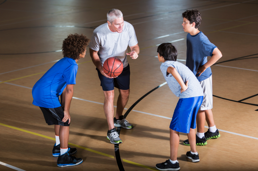 Ein Mann in Sportkleidung hält einen Basketball und erklärt etwas drei Jungen in einer Sporthalle. Die Jungen hören aufmerksam zu und sind ebenfalls sportlich gekleidet.