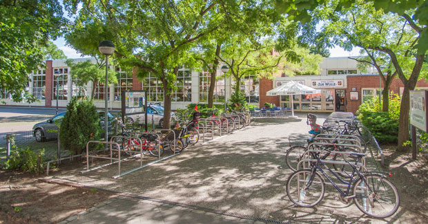EIngangsbereich des Zentrums: Ein Fahrradständer voller Fahrräder vor einem Gebäude mit roten Backsteinwänden und vielen Fenstern. Es gibt Bäume, die Schatten spenden, und einen Gehweg, der zum Eingang führt.