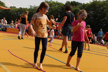 Kinder spielen draußen auf einem Spielplatz. Einige haben bunte Gesichter, und sie balancieren auf einem Seil am Boden. Im Hintergrund sind weitere Menschen zu sehen.