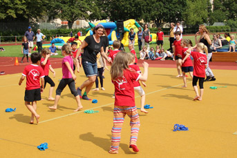 Kinder spielen barfuß auf einem Sportplatz. Sie tragen rote T-Shirts und werden von einem Erwachsenen betreut. Im Hintergrund sind Zuschauer zu sehen.