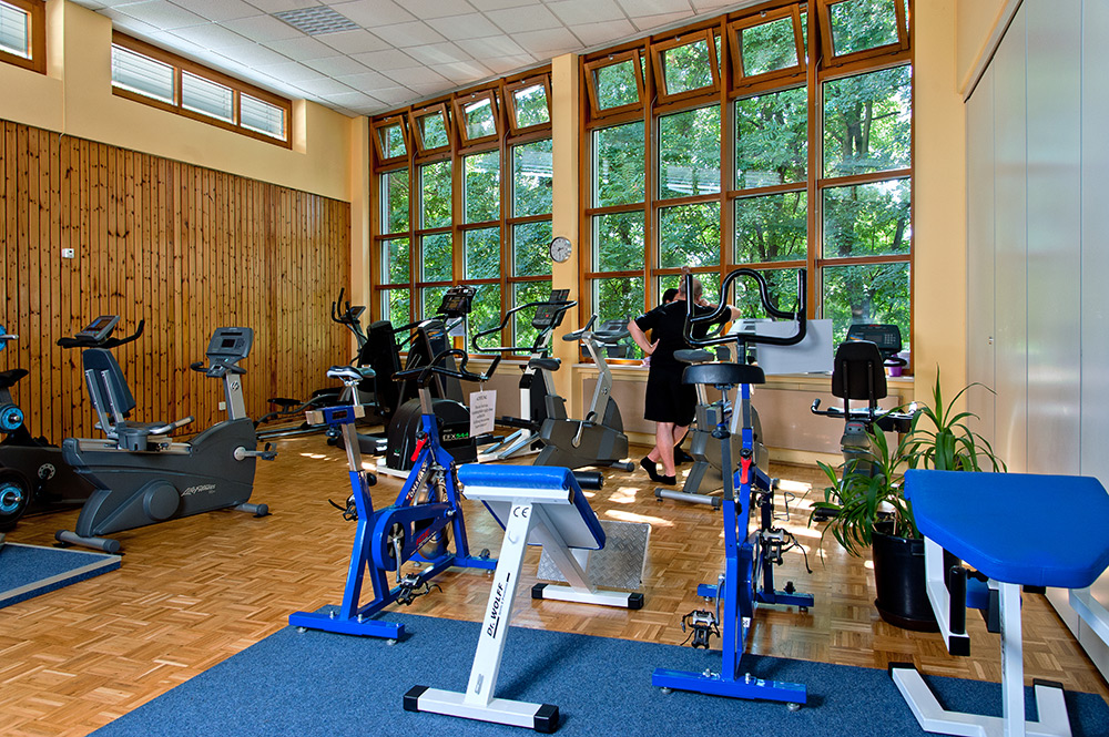 Ein Fitnessraum mit mehreren stationären Fahrrädern, einem Crosstrainer und einer Person, die an einem Gerät steht. Große Fenster bieten Blick ins Grüne. Der Boden ist aus Holz und teilweise mit blauen Teppichmatten bedeckt.