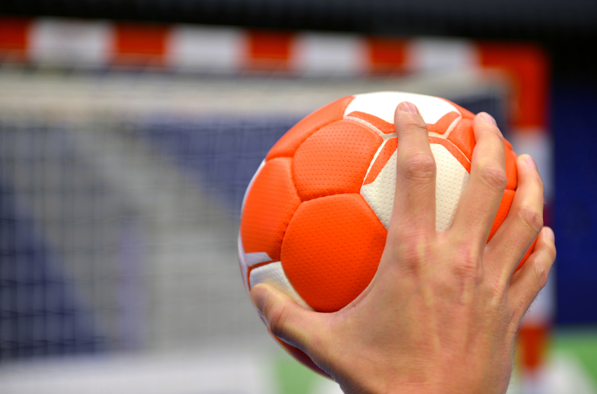 Ein Handball wird von einer Hand gehalten, mit einem unscharfen Tor im Hintergrund.