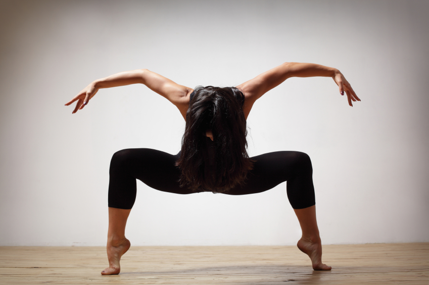 Eine Person in schwarzer Sportkleidung hält eine tief ausgefallene Pose mit gesenktem Kopf und ausgestreckten Armen in einem Raum mit Holzboden.
