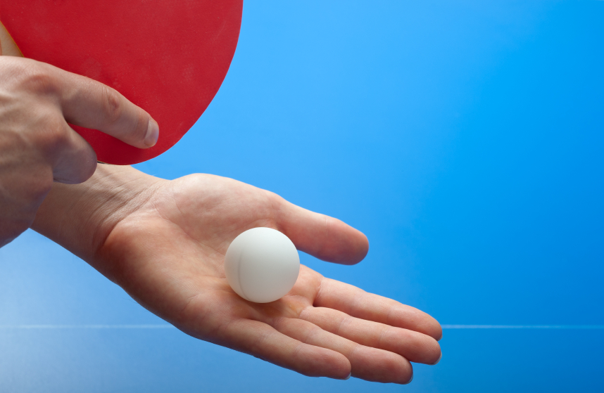 Eine Hand hält einen weißen Tischtennisball, während eine andere Hand einen roten Tischtennisschläger hält, vor einem blauen Hintergrund.