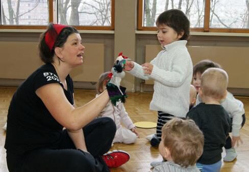 Eine Gruppe von Kleinkindern spielt mit einer erwachsenen Person in einem Raum mit Holzboden. Die erwachsene Person hält eine Handpuppe, während ein Kind freudig darauf zu reagiert. Im Hintergrund sind Fenster mit Blick nach draußen.