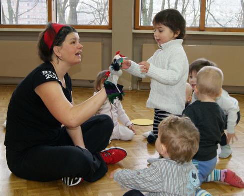 Eine Gruppe von Kleinkindern spielt mit einer erwachsenen Person in einem Raum mit Holzboden. Die erwachsene Person hält eine Handpuppe, während ein Kind freudig darauf zu reagiert. Im Hintergrund sind Fenster mit Blick nach draußen.