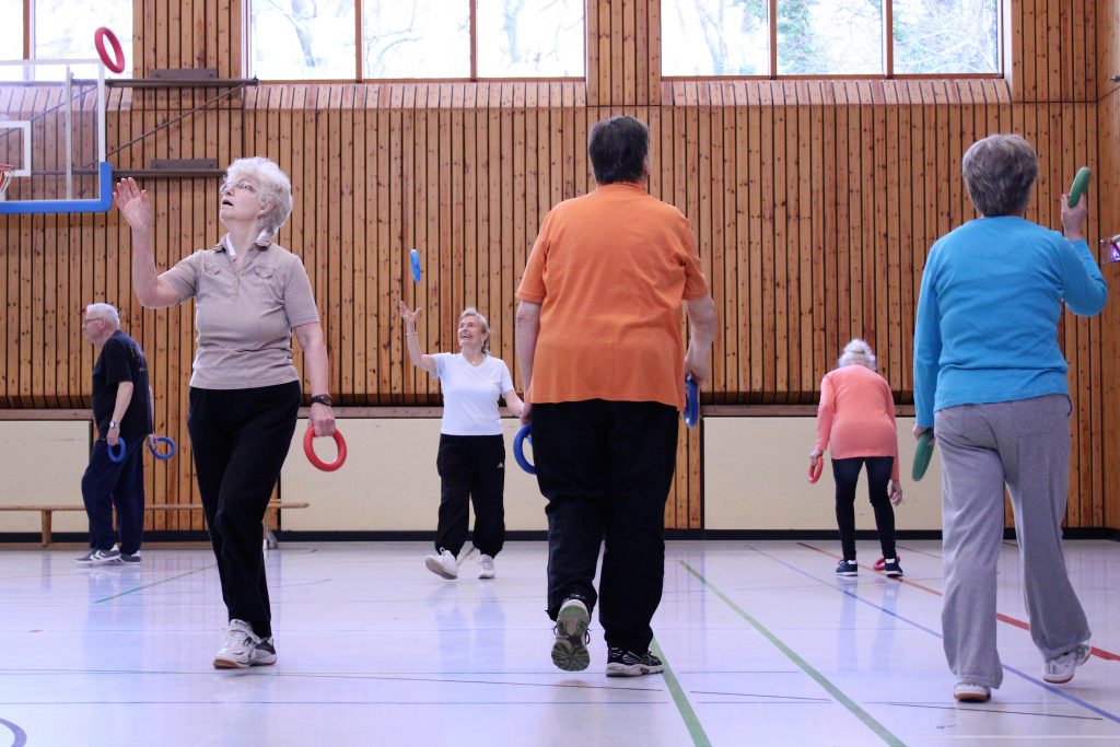 Ältere Menschen machen in einer Turnhalle gymnastische Übungen mit Ringen.