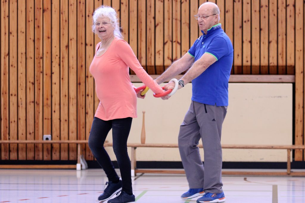 Ein älteres Paar tanzt in einer Sporthalle. Die Frau trägt ein oranges Oberteil und schwarze Leggings, während der Mann ein blaues Hemd und graue Hosen trägt. Beide halten einen roten Gymnastikstab.