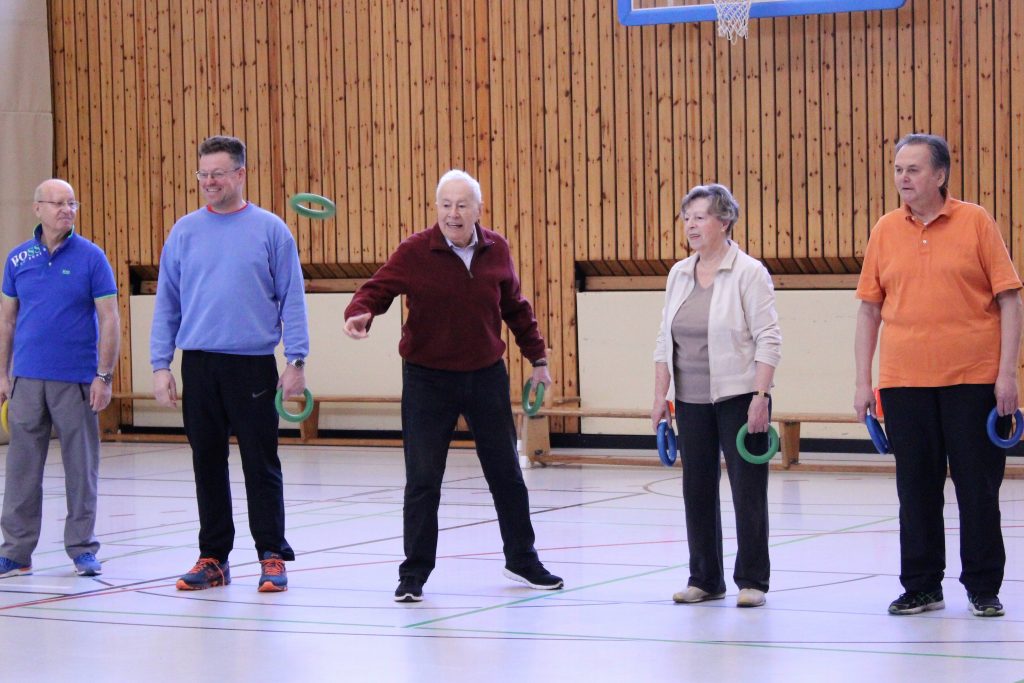Eine Gruppe von fünf älteren Personen steht in einer Sporthalle und hält grüne Ringe. Sie scheinen an einer Indoor-Aktivität oder Übung teilzunehmen.