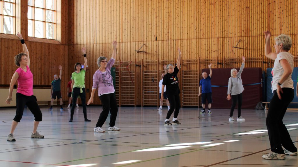 Eine Gruppe älterer Menschen macht Gymnastikübungen in einer Sporthalle. Alle heben einen Arm, während sie in Reihen stehen. Die Halle hat Holzverkleidungen und große Fenster lassen Licht herein.