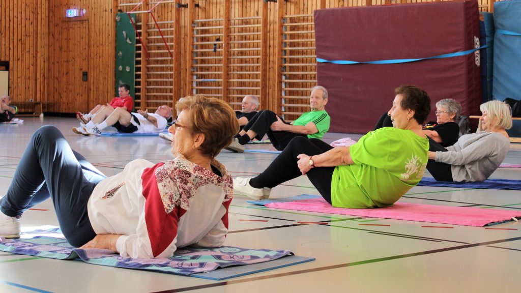 Ältere Erwachsene machen Gymnastikübungen auf Matten in einer Turnhalle. Einige tragen bunte T-Shirts, während sie auf dem Boden sitzen und sich zurücklehnen, um Übungen zu machen. Im Hintergrund sind Holzwände und Sprossenwände zu sehen.