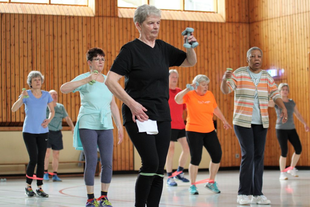 Eine Gruppe älterer Menschen macht Fitnessübungen in einer Turnhalle. Sie halten kleine Hanteln und haben Widerstandsbänder um die Knöchel. Die Teilnehmer tragen sportliche Kleidung.