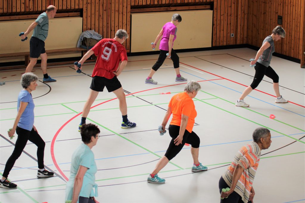 Ältere Menschen trainieren in einer Sporthalle mit Hanteln, während sie sich in Ausfallschritten bewegen. Sie tragen sportliche Kleidung und die Umgebung hat markierte Linien auf dem Boden.