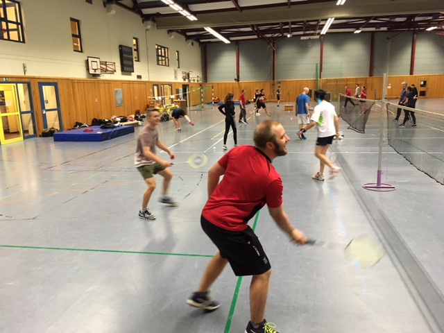 Mehrere Personen spielen Badminton in einer großen Sporthalle. Zwei Männer im Vordergrund spielen ein Doppelspiel, während andere im Hintergrund auf mehreren Plätzen aktiv sind.