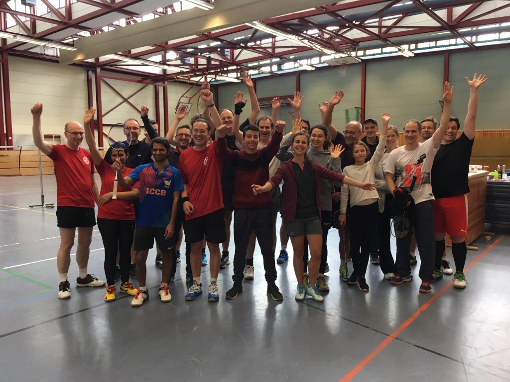 Eine Gruppe fröhlicher Menschen steht in einer Sporthalle, einige mit Badmintonschlägern in der Hand. Sie heben die Hände und lächeln in die Kamera.