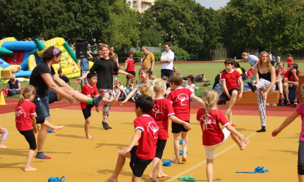 Eine Gruppe von Kindern und Erwachsenen macht Gymnastikübungen auf einem Sportplatz. Die Kinder tragen rote T-Shirts. Im Hintergrund ist eine bunte Hüpfburg zu sehen.