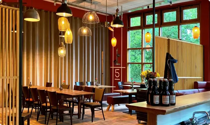 Wulle Café und Speisenwirtschaft - Innenansicht: Gemütliches Restaurant mit Holztischen und Holzstühlen, dekoriert mit verschiedenen hängenden Lampen. Große Fenster bieten Blick ins Freie.